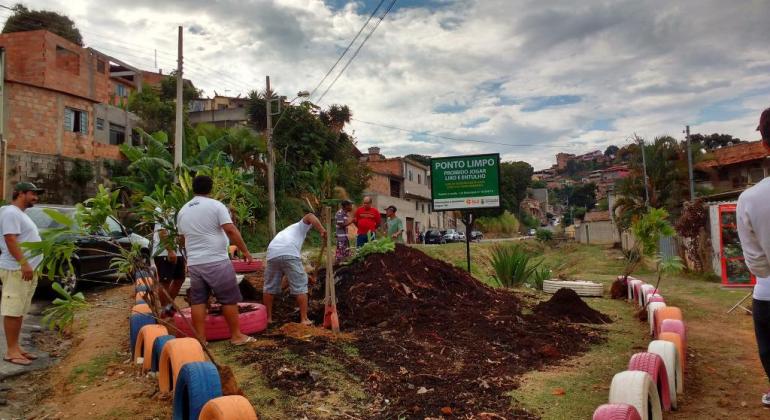 Imagem ilustrativa. Dois homens com instrumentos de capina cuidam de praça. Há, ao fundo, uma placa com texto "ponto limpo'