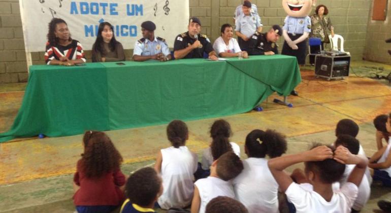 Durante seis meses, terceira edição do Projeto Adote um Músico ensina música a alunos do Alto Vera Cruz. Foto: Divulgação PBH Três membros da Guarda Municipal dividem a mesa de abertura do Projeto Adote um Músico com três membros da Escola Municipal Dom Orione; adiante, cerca de dez alunos assistem a abertura.