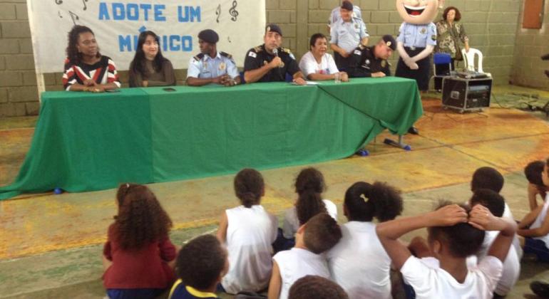 Três membros da Guarda Municipal dividem a mesa de abertura do Projeto Adote um Músico com três membros da Escola Municipal Dom Orione; adiante, cerca de dez alunos assistem a abertura. 