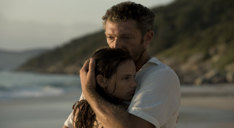 Homem abraça garota com praia ao fundo, durante o dia. Imagem do filme "À Deriva", em cartaz em mostra do MIS Cine Santa Teresa. 
