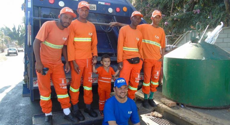 Garis da capital são admirados por crianças e adultos. Foto: Barbara Donadoni/PBH Criança ao lado de cinco profissionais da Limpeza Urbana na traseira do caminhão de lixo da SLU
