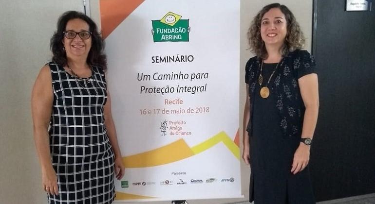 Duas mulheres ao lado de banner da Fundação Abrinq com os dizeres "Um Caminho para a Proteção Integral"