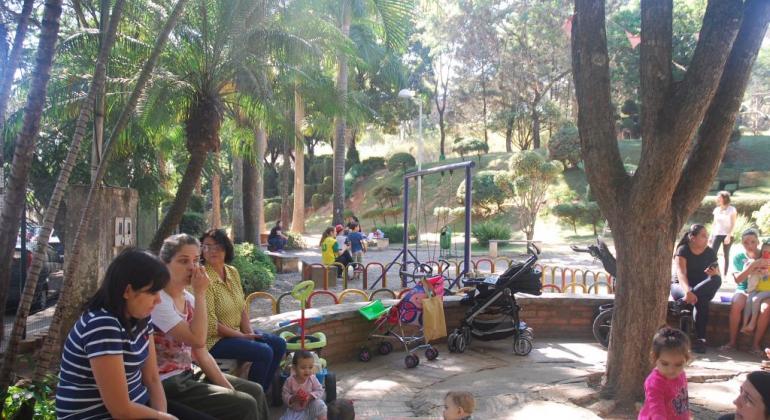 Crianças brincando no parque, mães ou responsáveis sentadas por perto. Todos debaixo de árvores. Ao fundo, o verde da vegetação do Parque.