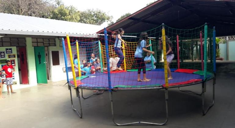 Programa Escola nas Férias oferece atividades diversificadas para toda a família. Foto: Divulgação/PBH Três crianças pulando em cama elástica no pátio da escola. Ao lado do brinquedo, outras crianças formam uma fila.
