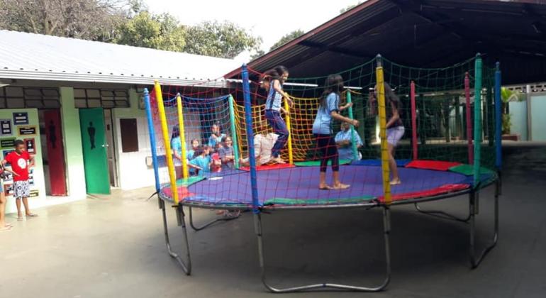 Três crianças pulando em cama elástica no pátio da escola. Ao lado do brinquedo, outras crianças formam uma fila.