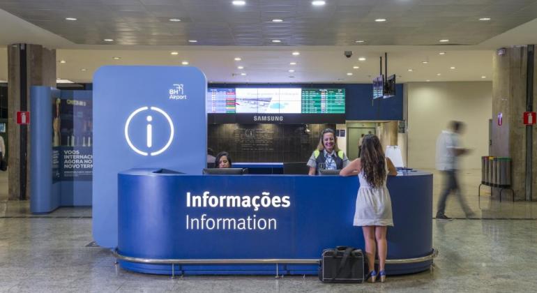 Belotur inicia pesquisa para identificar perfil do turista que visita a cidade Foto de balcão de informação azul no aeroporto de Confins. Há uma turista fazendo sua consulta.