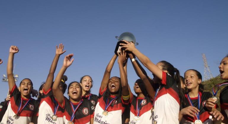 Equipe de futebol feminino sub-17 doTupinambás, todas as jogadoras com univorme e medalha, erguem a taça da Copa Centenário.