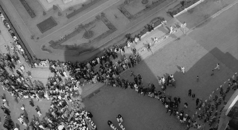 Foto histórica aérea em preto e branco de parte da cidade de Belo Horizonte, com mais de cinquenta pessoas, no tamanho de pontos pequeninos, a transitar pela rua. 