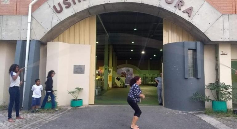 Oficina que resgata brincadeiras e brinquedos da infância é oferecida no Centro Cultural Nordeste. Foto: Melissa Reis Mulher pula corta na porta da Usina de Cultura, outras duas pessoas seguram e outras duas asssistem.