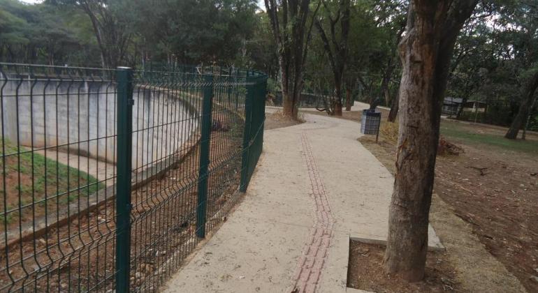 As obras de implantação de novas rotas acessíveis para pessoas com deficiência na Fundação Zoobotânica foram concluídas em julho. Foto: Antônio Rodrigues/PBH Passeio com marca de assessibilidade na Fundação Zoobotânica, ao lado de fosso para animais com grade, do outro lado, árvores.