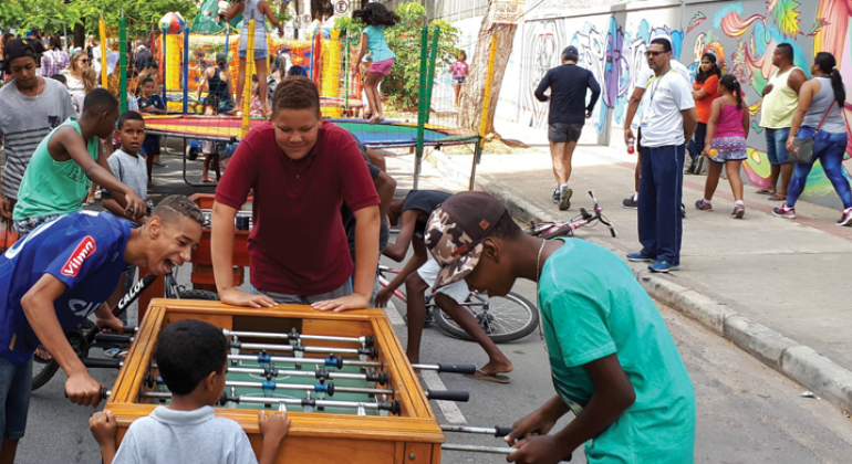 Atividades de recreação voltadas para o público infantil fazem parte da programação do BH é da Gente de domingo, dia 15/7. Foto: Vilma Tomaz Quatro meninos estão ao redor da mesa de pebolim na avenida Silva Lobo; atrás, dois outros brinquedos infantis e pessoas circulando, durante o dia.