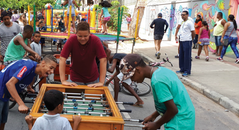 Quatro meninos estão ao redor da mesa de pebolim na avenida Silva Lobo; atrás, dois outros brinquedos infantis e pessoas circulando, durante o dia. 