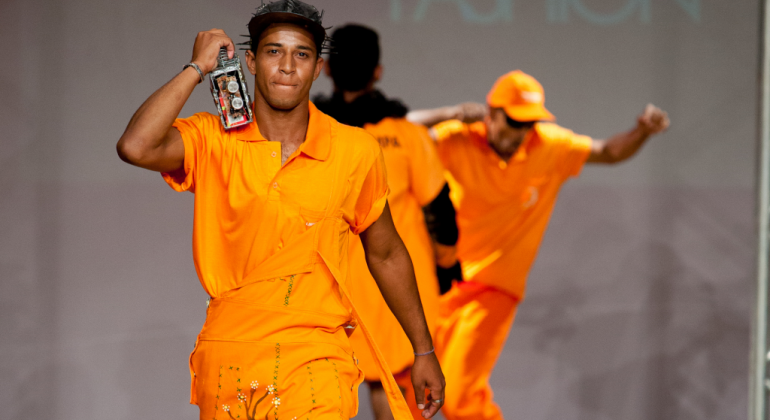 Garis desfilam em passarela do Gari Fashion. Dois deles estão de frente e um de costas. Todos eles usam laranja.