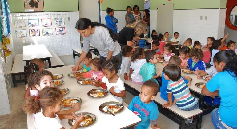 Mais de vinte crianças de 3 anos de idade almoçam, sob a supervisão de profissional da Secretaria Municipal de Educação. 