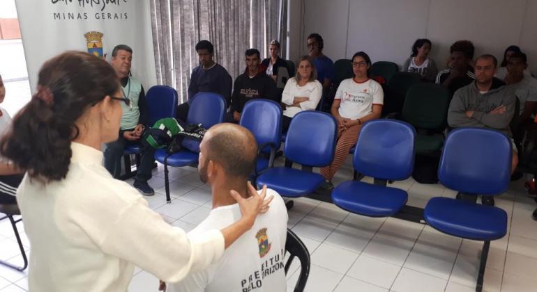 Estagiários do programa Vida Ativa participam de capacitação A foto mostra uma estagiária do Programa Vida Ativa realizando um treinamento em um homem que está sentado em uma cadeira, enquanto outros estagiários assistem a atividade.