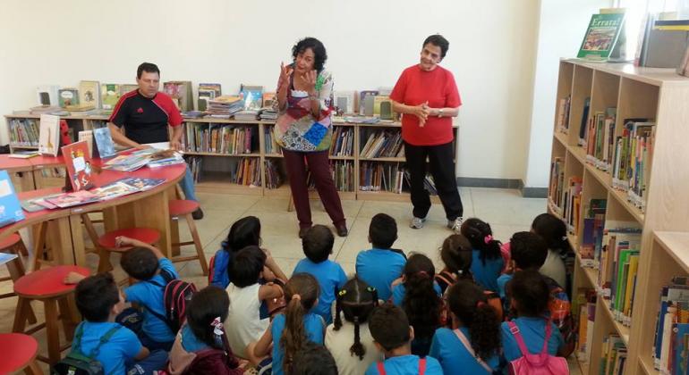 Cerca de vinte crianças sentadas no chão enquanto três adultos contam e interpretam histórias em uma biblioteca.