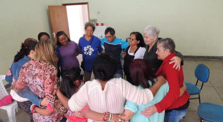 Grupo de cerca de quinze pessoas abraçados formando uma roda dentro de uma sala