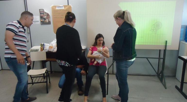 Formação de profissionais das unidades básicas de saúde favorece amamaentação. Foto: Avanilton de Aguiar/PBH Pais participam de oficina de amamentação acompanhados de equipe de profissionais da saúde. Mulher segurando boneca, acompanhada do marido e três outras pessoas.