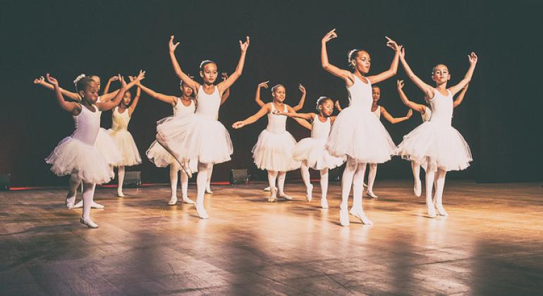  A foto mostra dez bailarinas usando colant e saia tu-tu brancos, em pose típica de bailarinas em um palco. 