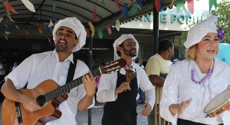 três artistas com diferentes instrumentos: Chocalho, pandeiro e vioão e caracterizados como cozinheiros em frente ao Restaurante Popular