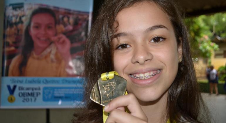 Garota segura medalha da OBMEP; ao fundo, Banner da Obmep com foto da mesma garota.