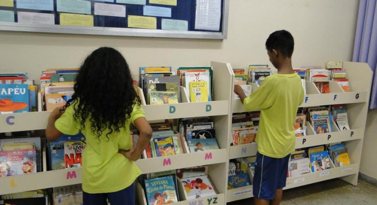 Programa Leituras em Conexão promove o interesse pela leitura e escrita. Foto: Divulgação/PBH Duas crianças na biblioteca escolar escolhendo livros na prateleira