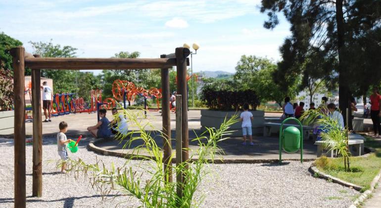 Parque com equipamentos de ginástica, brinquedos, área de convivência, lixeira, vegetação variada, crianças brincando e adultos praticando diversas atividades.