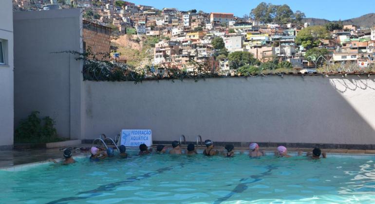 Treze crianças em piscina praticando natação; ao fundo, aglomerado da Serra.