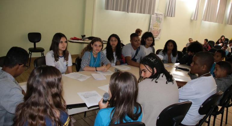 Cerca de quinze jovens alunos da Rede Municipal sentados em uma mesa com microfones e documentos.