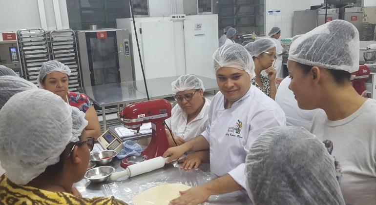 Vila Viva promove curso de culinária para mulheres do Morro das Pedras. Foto: Divulgação/PBH Cerca de quinze mulheres, e professor, na cozinha manuseando massas e aperfeiçoando sua culinária