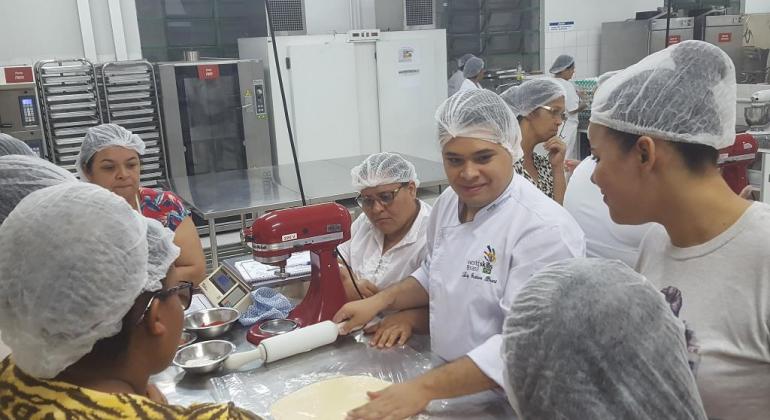 Cerca de quinze mulheres, e professor, na cozinha manuseando massas e aperfeiçoando sua culinária