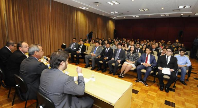 De costas, quatro homens sentados, dois deles são o Prefeito em exercício, Paulo Lamac, e o Procurador Geral do Município. De frente, uma plateia de cerca de cinquenta pessoas. 