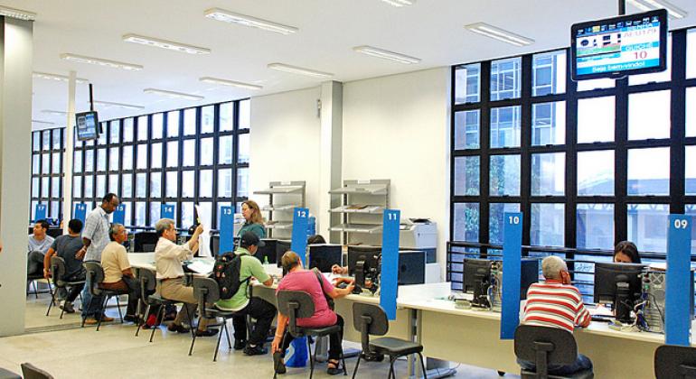 A foto mostra a sala de atendimento presencial do BH Resolve. Há oito pessoas sentadas em guichês, conversando com os atendentes do outro lado da mesa. 