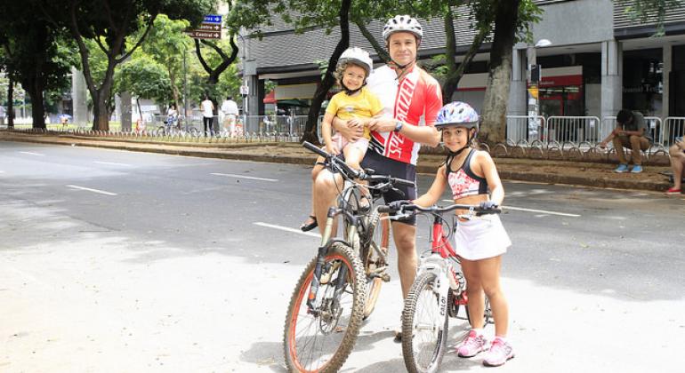 A foto mostra um adulto e duas crianças com bicicletas. Eles usam capacete e equipamentos de segurança. Uma das crianças está no colo do adulto.