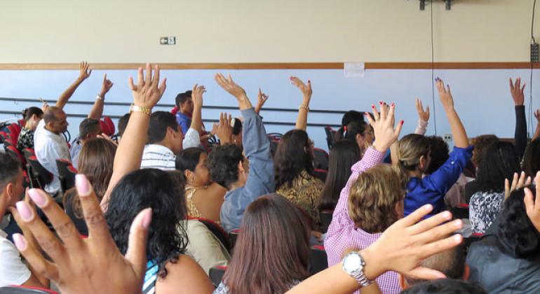 A imagem mostra cerca de 30 pessoas sentadas em um auditório, com as mão levantadas, em uma votação.