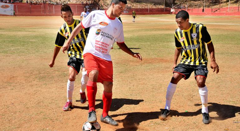 Na foto há três jogadores de futebol disputando uma bola. Dois usam uniforme preto e amarelo e um usa uniforme branco e vermelho.