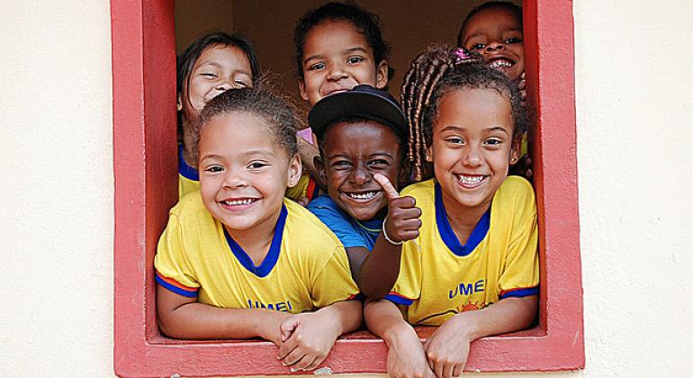 A foto mostra seis crianças, de quatro e cinco anos, em uma janela, sorrindo para a câmera.