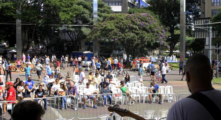 Em palco montado na Savassi, músicos estão de costas se apresentando e a platéia, sentada no meio da Praça, assiste ao show.