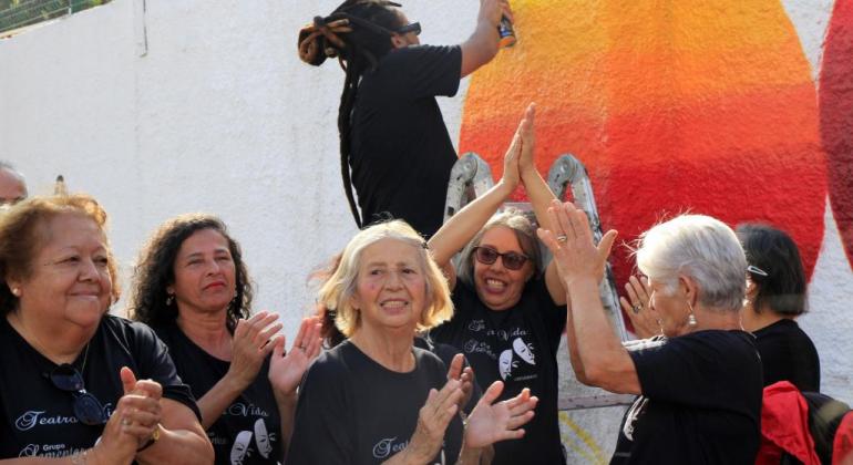 Na foto há um artista grafitando o muro do Centro de Referência da Pessoa Idosa. No primeiro plano, seis idosos batem palmas. 