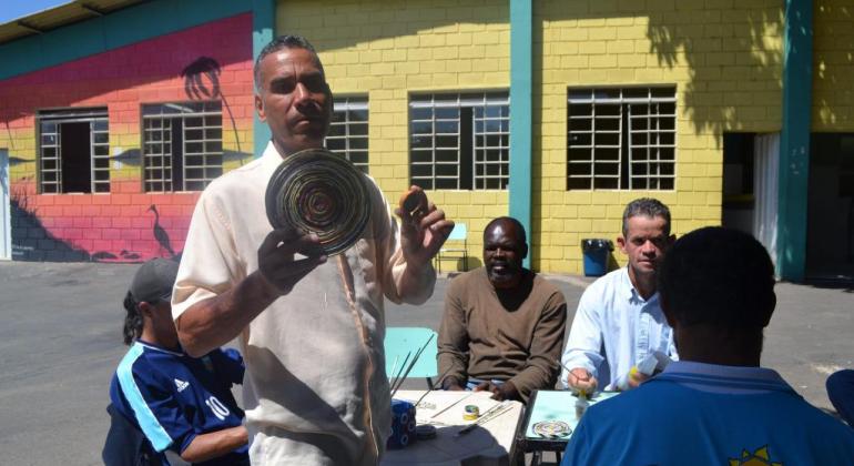 Um homem segurando peça de artesanato e outros quatro sentados produzindo durante oficina