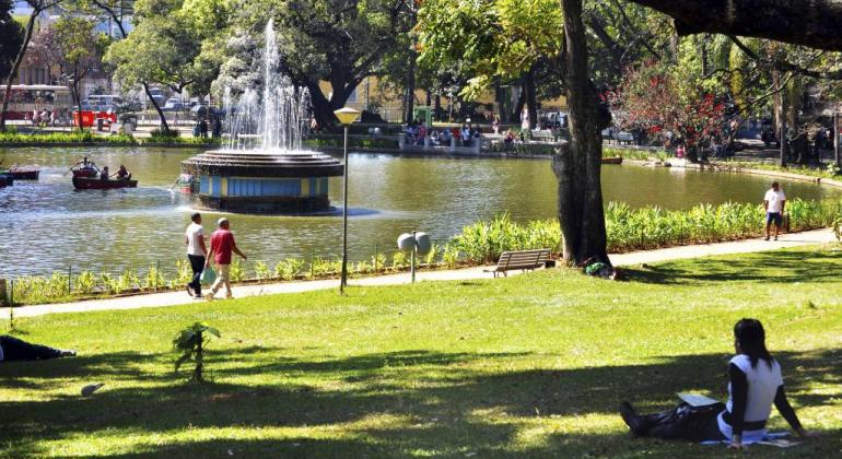 Cidadãos caminhando na orla do lago e uma mulher descansa sob as árvores no Parque Municipal Américo Renné Giannetti
