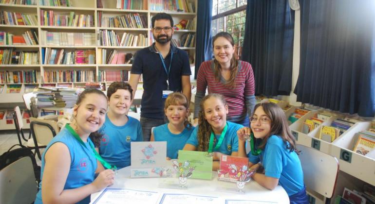 Dois profissionais da educação e cinco crianças na biblioteca exibindo certificados de premiação e medalhas