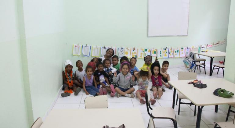 Professora da educação infantil e dezenas de crianças da creche em sala de aula, sentados no chão.