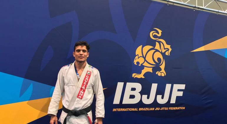 Homem de quimono ao lado de banner da IBJJF, Federação Internacional de Jiu-Jitsu Brasileiro
