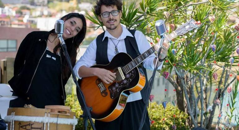 A foto mostra dois músicos em apresentação. À esquerda, uma mulher toca um tambor e, à direita, um homem toca violão.