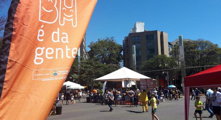 Praça da Savassi, fechada para os carros, com barracas e pessoas caminhando. À esquerda, um banner escrito BH é da Gente.