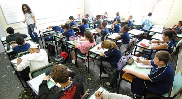 Foto mostra sala de aula com alunos com idade aproximada de 11 anos, sentados em carteiras. Na frente da sala há uma professora em pé, próxima ao quadro branco.