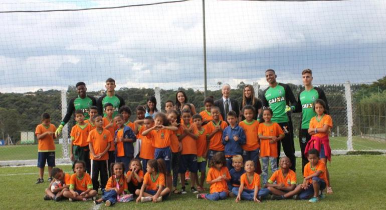 Crianças com camisas na cor laranja do projeto escola integrada posam com quatro goleiros do time do América