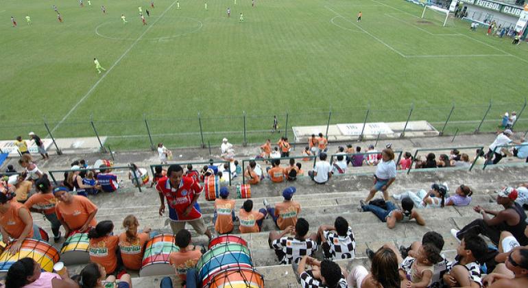 A foto mostra a vista de um campo de futebol a partir da arquibancada, onde é possível ver cerca de cinquenta pessoas