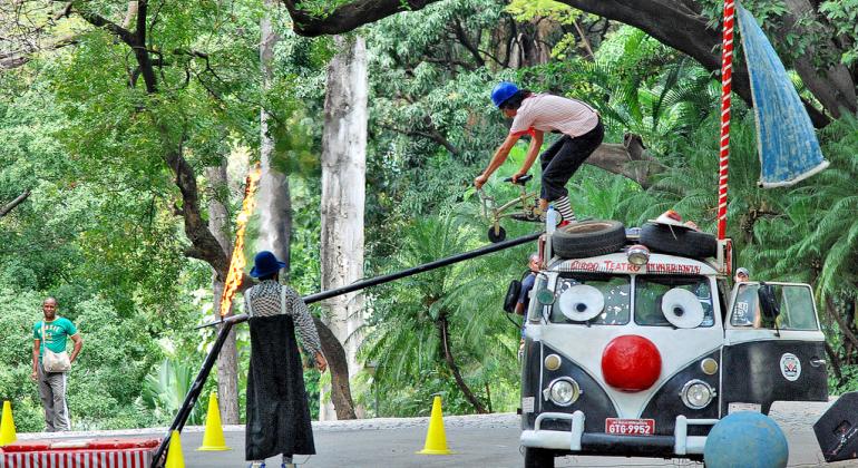 A foto mostra atores em uma apresentação de teatro no Parque Municipal. No canto direito da imagem há uma kombi colorida em alusão a um palhaço.